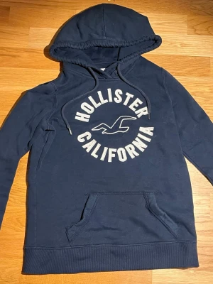 Mörkblå hoodie från Hollister XS - Mörkblå hoodie från Hollister med stor vit logga och texten 'Hollister California' på bröstet. Klassisk känguruficka framtill, dragsko i huvan och ribbade muddar. Perfekt för en avslappnad och cool stil. Den passar dig som är mellan 160-170