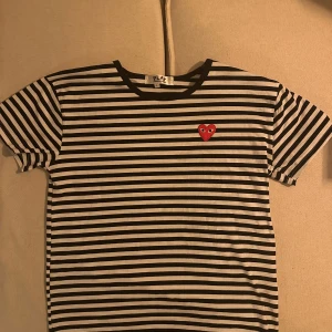 Randig t-shirt från Comme des Garçons - Säljer en svartvit randig t-shirt från Comme des Garçons Play med det ikoniska röda hjärtat broderat på bröstet. T-shirten har korta ärmar och rund halsringning. Perfekt för dig som gillar streetstyle och vill sticka ut med en klassisk design.