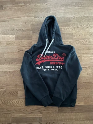 Superdry hoodie - Svart hoodie från Superdry i storlek L med stor röd och vit logga på bröstet samt japansk text. Klassisk huva med vita dragsnören. Tröjan har vissa skavanker men är fortfarande i bra skick. Tröjan har även en QR-kod (se på bild 3).           Skriv vid frågor, pris kan diskuteras 🤝