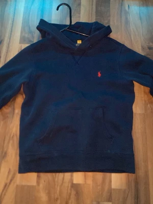 Mörkblå hoodie från Polo Ralph Lauren - Mörkblå hoodie från Polo Ralph Lauren med klassisk röd logga broderad på bröstet. Hoodien har en stor känguruficka framtill och en skön huva. Tillverkad i mjukt bomullsmaterial som känns riktigt bekvämt. Perfekt för en avslappnad och stilren look. !! Och en hål  på armbågen , går att sy ihop