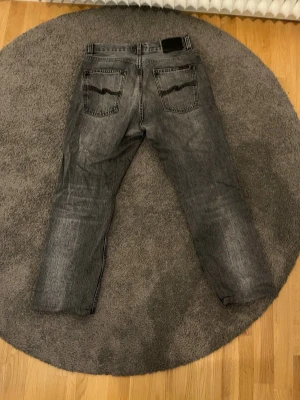 Svarta jeans från Nudie Jeans - Säljer ett par svarta jeans från Nudie Jeans med klassisk femficksdesign och raka ben. Jeansen har en snygg tvättad look och kontrastsömmar på bakfickorna. Perfekta för en avslappnad stil och tillverkade i bomull.