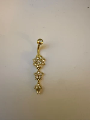 Guldfärgad navelpiercing med stenar - Säljer en snygg guldfärgad navelpiercing med flera små gnistrande stenar i blommönster. Piercingen har en böjd stav och hängande dekorationer som ger ett elegant intryck. 