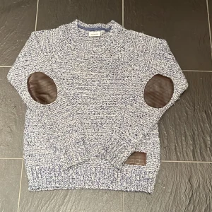 Blåvit stickad tröja med mockadetaljer - Säljer en blåvit melerad stickad tröja från Kappahl med bruna mockalappar på armbågarna och en detalj vid nederkanten. Tröjan har rund hals, långa ärmar och ribbstickade muddar. Perfekt för dig som gillar klassisk men trendig stil.