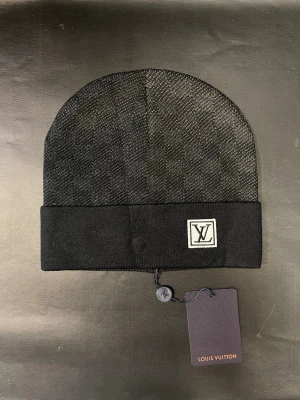 Svart mössa med logga - Snygg svart mössa med diskret rutmönster och vikt kant. På framsidan sitter en vit patch. Mössan är stickad och har en exklusiv känsla, perfekt för dig som gillar stilrena accessoarer.