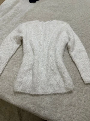 Vit fluffig stickad tröja - Mysig vit stickad tröja med fluffig textur och lång ärm. Tröjan har en smal passform och är perfekt för kalla dagar. Materialet ser ut att vara en mjuk syntetblandning som ger en skön känsla mot huden.