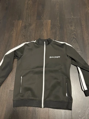Palm Angels svart track jacket - Svart track jacket från Palm Angels med vita ränder längs ärmarna och vit dragkedja framtill. Jackan har ståkrage, två fickor med dragkedja och logga på bröstet. Perfekt för en sportig och trendig look.