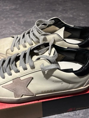 Golden Goose vita sneakers med stjärna - Snygga vita sneakers från Golden Goose med grå mockastjärna på sidan, grå skosnören och svart hälparti. Skorna har en klassisk låg siluett och är tillverkade i läder med detaljer i mocka. Perfekta för dig som gillar streetstyle och unika detaljer.