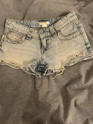 Ljusblå jeansshorts med broderi - Snygga ljusblå jeansshorts med slitna kanter och broderade detaljer på bakfickorna. Shortsen har låg midja och klassisk femficksdesign. Perfekta för varma dagar och en avslappnad stil.