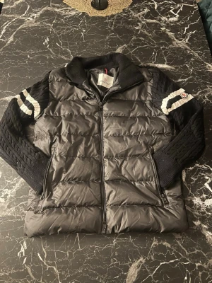 Moncler cardigan knitted - Säljer en svart dunväst från Moncler med stickade ärmar och detaljer i vitt på armarna. Västen har dragkedja framtill och quiltad framsida i glansigt material, medan baksidan och ärmarna är grovstickade. Perfekt för dig som vill ha en sportig och stilren look.