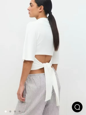 Vit croppad topp med knyt i ryggen - Säljer en vit croppad topp med halvlånga ärmar och snygg knytning i ryggen som ger öppen rygg. Toppen har en enkel och clean look, perfekt till lounge eller chill outfits. Materialet ser ut att vara mjukt och stretchigt, troligen bomull eller en bomullsblandning.