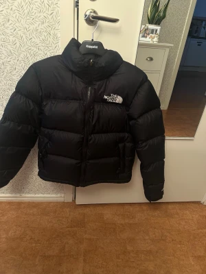 Svart pufferjacka från The North Face - The north face retro Nuptse -dunjacka i nyskick, endast använd ca 3-4 gånger. Det finns även en luva på jackan som är ”instoppad” för att döljas när man vill det. 