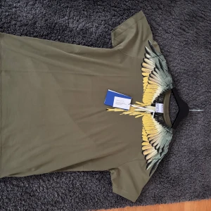 Olivgrön t-shirt med vingtryck från Marcelo Burlon - Cool olivgrön t-shirt från Marcelo Burlon med unikt vingmönster i gult, svart och grönt över axlar och bröst. T-shirten har rund hals och korta ärmar. Perfekt för dig som vill sticka ut med en statement-piece.