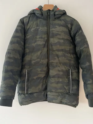 Dunjacka med huva i kamouflage - Snygg dunjacka i grönt kamouflagemönster med dragkedja framtill och två fickor med reflexdetaljer. Jackan har en stor huva och ribbade muddar vid ärmsluten. Perfekt för kalla vinterdagar och ger en cool streetwear-look.