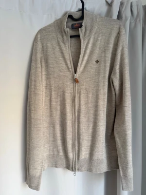 Beige ziptröja från Morris - Snygg beige ziptröja från Morris med diskret logga broderad på bröstet. Tröjan har hög krage, tvåvägsdragkedja och ribbade muddar vid ärmslut och nederkant. Perfekt att slänga på sig över en t-shirt för en clean look.