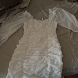 Vit kort klänning med puffärmar - Supersöt vit kort klänning med puffiga, transparenta långärmar och rynkade detaljer framtill. Klänningen har en v-ringad urringning och figurnära passform. Perfekt för studenten eller andra somriga tillfällen!!