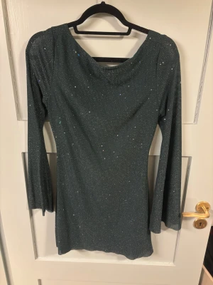 Glittrig mörkgrön klänning med öppen rygg - Snygg mörkgrön kortklänning med glittrigt tyg och långa ärmar. Klänningen har en djup öppen rygg med knytband och en lätt draperad ringning både fram och bak. Perfekt för dig som vill sticka ut med en elegant och trendig look.