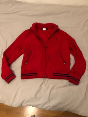 Ralph Lauren Salesman Sample – Spring 2008 - Exclusive because of its origin, not because of marketing.  Röd lätt vadderad jacka med dragkedja från Polo Ralph Lauren. Detta är en Salesman Sample, vilket innebär att jackan inte var avsedd för vanlig butikförsäljning utan användes i showroom/av säljsrepresentanter. Producerad i mycket begränsat antal.