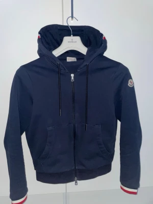 Marinblå Moncler kofta - Säljer en tvär snygg Moncler kofta som är köpt för 5 000 kr 2024. Tar inte på mig fejk! Den är väldigt sparsamt använd och alltid använd försiktigt, så den är i superfint skick.  Koftan sitter riktigt bra och funkar till asså fester, restauranger och i skolan. Varje gång jag haft på mig den har jag fått komplimanger, så den sticker definitivt ut så att säga. Hör av dig vid minsta lilla fundering. 
