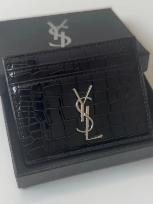 YSL korthållare i krokodilpräglat läder - Snygg svart korthållare från Yves Saint Laurent i glansigt läder med krokodilpräglad yta. Framsidan har det ikoniska YSL-logotypen i silver. Perfekt för kort och enkel att ha i fickan. Helt oanvänd (ny). Kan gå ner i pris vid snabb affär. Skriv vid minsta lilla fundering!