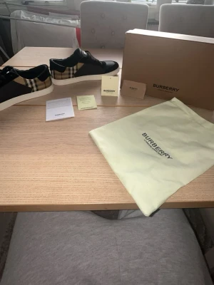 Burberry skor  - Snygga låga sneakers från Burberry i svart läder med klassiskt beige rutmönster på sidan och vita sulor. Skorna har svarta snören och Burberry-logga på plösen. Perfekta för dig som vill ha en clean och lyxig stil.