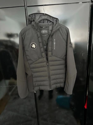 Sail racing jacka - Säljer en svart dunjacka från Sail racing med huva och dragkedja framtill. Jackan har quiltad design på fram- och baksida, samt två märken på bröstet. Tillverkad i syntetmaterial och polyester, perfekt för kalla dagar.