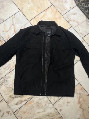 Svart mocka jacka från Only & Sons - Stilren svart mocka jacka från Only & Sons med dragkedja framtill och två bröstfickor. Jackan har klassisk krage och raka ärmar, perfekt för lager-på-lager. Materialet är mjukt och lätt, vilket gör den enkel att bära till olika outfits. Köpt ny i höstas kommit till användning väldigt få gånger.