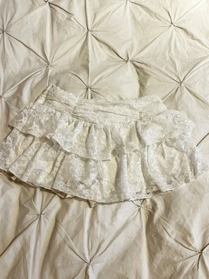 Vit spetsig skort med volanger - Supersöt vit skort med dubbla volanger i spets och inbyggda shorts. Perfekt för dig som vill ha kjol-looken men ändå känna dig bekväm. Spetsen har blommigt mönster och midjan är elastisk för extra komfort.