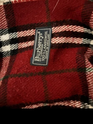 Rutig vinröd halsduk från Burberrys - Klassisk halsduk från Burberrys i vinrött med rutigt mönster i svart, vitt och beige. Tillverkad i 100% lammull och har fransar i ändarna. Perfekt accessoar för att lyfta din outfit med en tidlös brittisk stil.