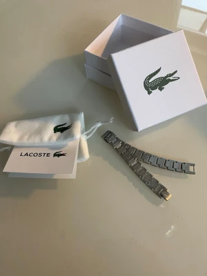 Silverfärgat armband från Lacoste - Snyggt armband i silverfärgad metall från Lacoste med det klassiska krokodilmärket ingraverat på spännet. Länkarna har en cool, strukturerad design och armbandet kommer i originalask och tygpåse med logga.
