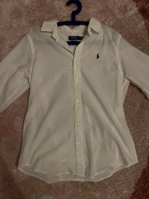 Vit skjorta från Polo Ralph Lauren - Klassisk vit skjorta från Polo Ralph Lauren med blå broderad logga på bröstet. Skjortan har lång ärm, knappar framtill och en tidlös krage. Perfekt till jeans eller chinos för en clean och stilren look.