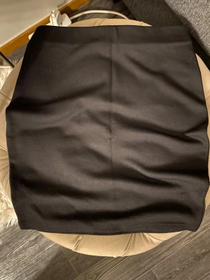 Svart tight kortkjol - En enkel och stilren svart kortkjol med tight passform. Kjolen har en rak och klassisk design utan synliga detaljer eller mönster, perfekt att matcha med olika toppar och jackor.