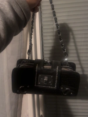 Svart handväska med kedjerem - Snygg svart handväska med silverdetaljer och kedjerem. Väskan har ett rektangulärt format, dekorativa nitar och krokodilpräglat mönster. Framtill finns ett spänne och dragkedja, vilket ger en edgy look. Perfekt accessoar för att lyfta din outfit.