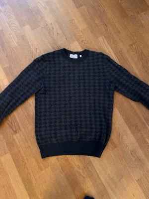 Mörkgrå stickad tröja med hundtandsmönster från Bläck - Snygg mörkgrå stickad tröja med klassiskt hundtandsmönster. Tröjan har rund halsringning, långa ärmar och ribbade muddar vid ärmslut och nederkant. Perfekt för dig som gillar stilrena och tidlösa plagg.