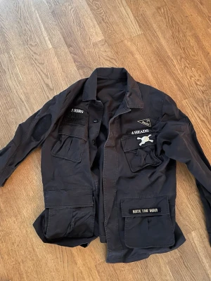 Svart overshirt med patches - Cool svart overshirt med flera fickor och unika patches som '3 RIBS', '4 HEADS' och 'HATE THE WAR'. Overshirten har knäppning framtill och detaljerade broderier på bröstet. Perfekt för dig som gillar streetwear och statement-plagg.