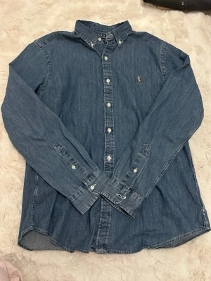 Blå jeansskjorta från Polo Ralph Lauren - Snygg blå jeansskjorta från Polo Ralph Lauren med klassisk button-down krage och broderad logga på bröstet. Slim fit-modell med långa ärmar och vita knappar. Perfekt för en avslappnad men stilren look.