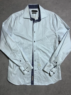 Vit smalrandig skjorta från Riley - Vit skjorta från Riley med tunna ljusblå ränder och kontrastmönstrad insida i krage och knappslå. Slim fit-modell med långa ärmar och klassisk krage. Perfekt för dig som gillar stilrena detaljer och snygg passform. Den har en gul fläck på bilden därav priset 