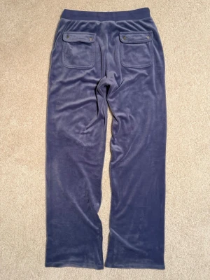 Dimblå Juicy Contoure mjukisbyxor med fickor. - Juicy Countore byxor i mjuk velour med raka ben och dragsko i midjan. Byxorna har stora fickor fram och bak, bakfickorna har lock och knapp.  Foll längst ner på vänster ben har gått upp lite, inget som märks(se bild)Nypris 1000kr. Storlek 12-13 år, cirka 158cl. 