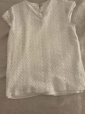 Vit spetsblus från Only - Söt vit blus från Lindex med korta ärmar och rund halsringning. Blusen är täckt av vacker spets med blommigt mönster och har knäppning bak i nacken. Perfekt att styla med jeans eller kjol för en fräsch look.