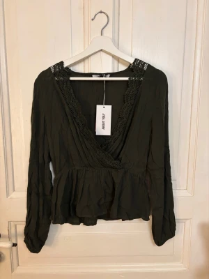 Olivgrön omlottblus med spetsdetaljer - Trendig olivgrön omlottblus från ABOUT YOU med långa ärmar och vacker spets längs den djupa v-ringningen. Blusen har volang nertill och är tillverkad i ett lätt, luftigt tyg som ger en avslappnad men chic look.