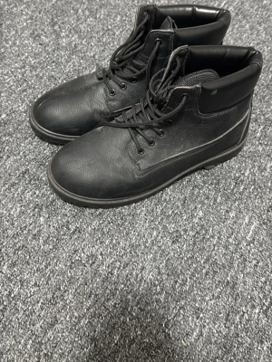 Svarta höga kängor i läderlook - Snygga svarta kängor med högt skaft och snörning framtill. Kängorna har en stilren design med vadderad kant upptill och detaljer i läderimitation. Perfekta för dig som gillar en tuff och urban stil.