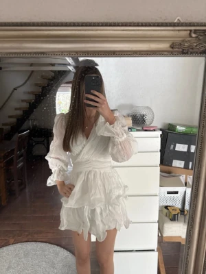 Vit omlottklänning med volanger - Supersöt vit kort omlottklänning med volanger och långa puffärmar. Den är super fin och skön men känns inte som en klänning för mig. Passar perfekt till studenten!