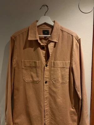 Beige overshirt från H&M - Stilren beige overshirt från H&M med två stora bröstfickor och klassisk krage. Tillverkad i ett kraftigare bomullstyg och har svarta knappar framtill. Perfekt att bära öppen över en t-shirt för en avslappnad look.