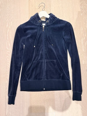 Mörkblå velourhoodie med dragkedja - Mysig mörkblå hoodie i mjuk velour med dragkedja framtill och två stora fickor. Justerbar huva med snören och ribbade muddar vid ärmslut och nederkant. Perfekt för en avslappnad och trendig stil.