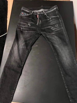 Svarta slim fit jeans med slitningar - Säljer ett par svarta slim fit jeans med coola slitningar framtill och på bakfickorna. Jeansen har klassisk femficksdesign, dragkedjegylf och detaljer i rött vid fickan. Perfekta för en trendig streetwear-look.