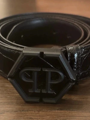 Svart skärp från Philipp Plein - Snyggt svart skärp från Philipp Plein med en unik sexkantig spänne och PP-logga framtill. Skärpet är gjort i präglat skinn med en glansig finish som ger en lyxig känsla. Perfekt accessoar för att lyfta din outfit.