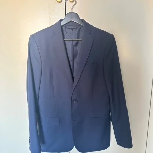 Mörkblå kavaj från J.Lindeberg - Snygg mörkblå kavaj från J.Lindeberg i slim fit-modell. Klassisk design med två fickor framtill, bröstficka och enkla knappar vid ärmslut. Perfekt för en stilren look. Materialet är slätt och har en lätt glansig finish. Bra skick! Pris:499