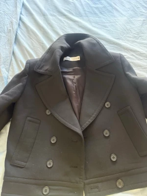 Svart skepparkavaj från Zara - Stilren svart skepparkavaj från Zara med dubbelknäppning och breda slag. Kavajen har två raka fickor framtill och är helfodrad för extra komfort. Klassisk och tidlös modell som passar perfekt till höst och vinter.