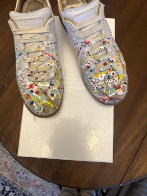 Maison Margiela sneakers med färgstänk - Unika vita sneakers från Maison Margiela med färgstänk i rött, blått, gult och svart. Klassisk låg modell med snörning och läderdetaljer. Snygg och konstnärlig design som sticker ut i mängden. Äkta.