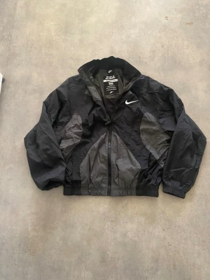 Svart vindjacka från Nike - Retroinspirerad svart vindjacka från Nike med grå detaljer och klassisk vit swoosh-logga på bröstet. Jackan har dragkedja, resår i ärmslut och midja samt meshfoder. Perfekt för en sportig och stilren look.