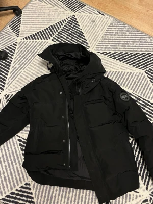 Svart dunjacka från Canada Goose - Säljer en svart dunjacka från Canada Goose med klassisk patch på ärmen. Jackan har huva, dragkedja och knappar framtill samt flera fickor. Perfekt för kalla vinterdagar och riktigt varm tack vare dunfyllningen.